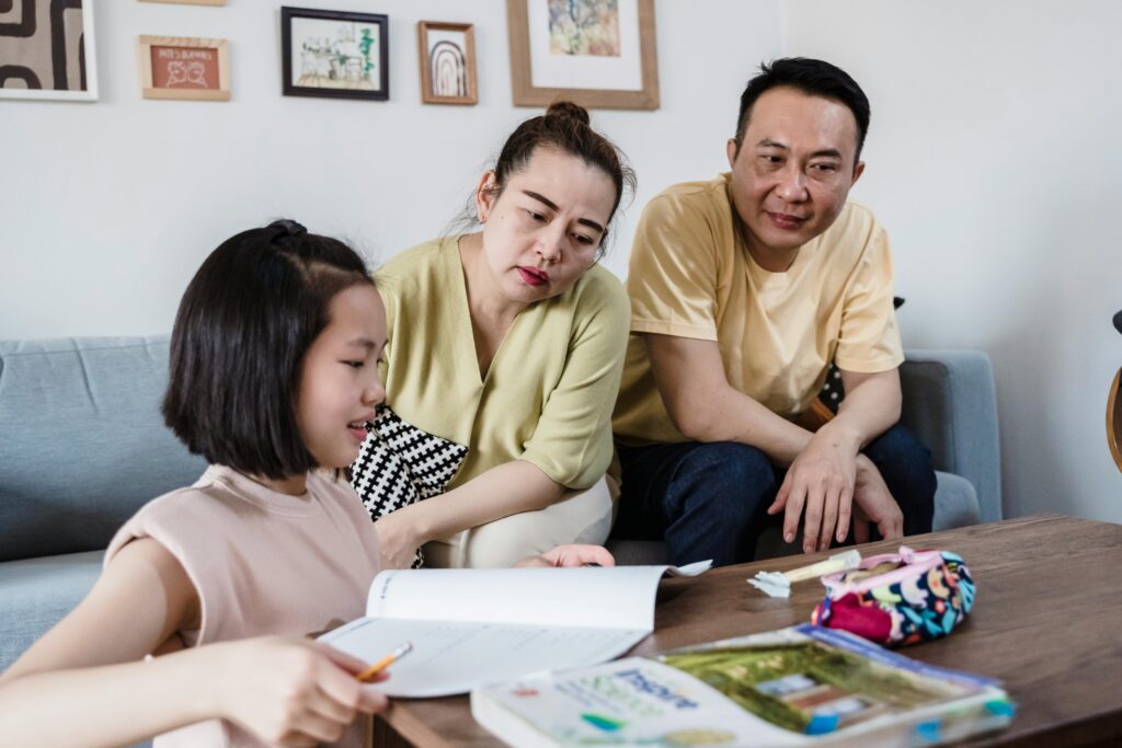 A young girl does homework at a table while two adults sit on a sofa behind her, watching and smiling. | TCS