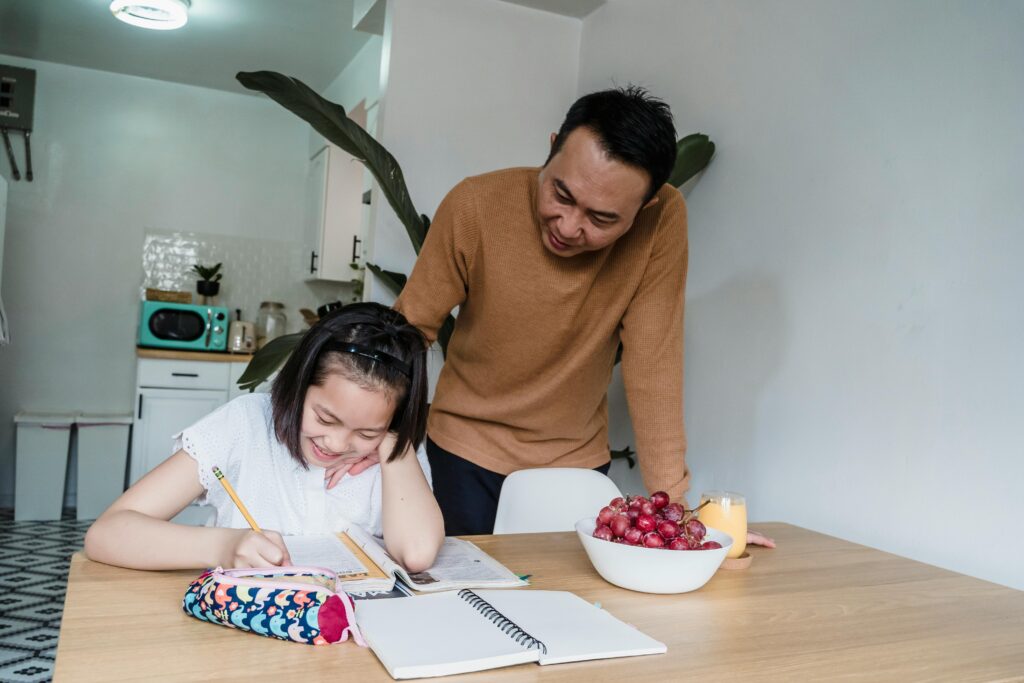 A man smiles as he helps a young girl with her homework at a table with fruit and a glass of juice. | TCS