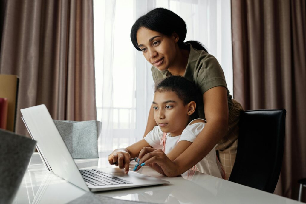 A woman helps a young girl use a laptop at a desk in a bright room with curtains. | TCS