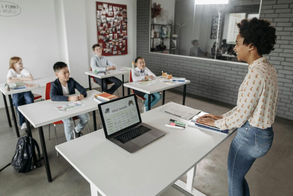 A teacher stands at the front of a classroom, smiling at four seated pupils with a laptop on her desk. | TCS