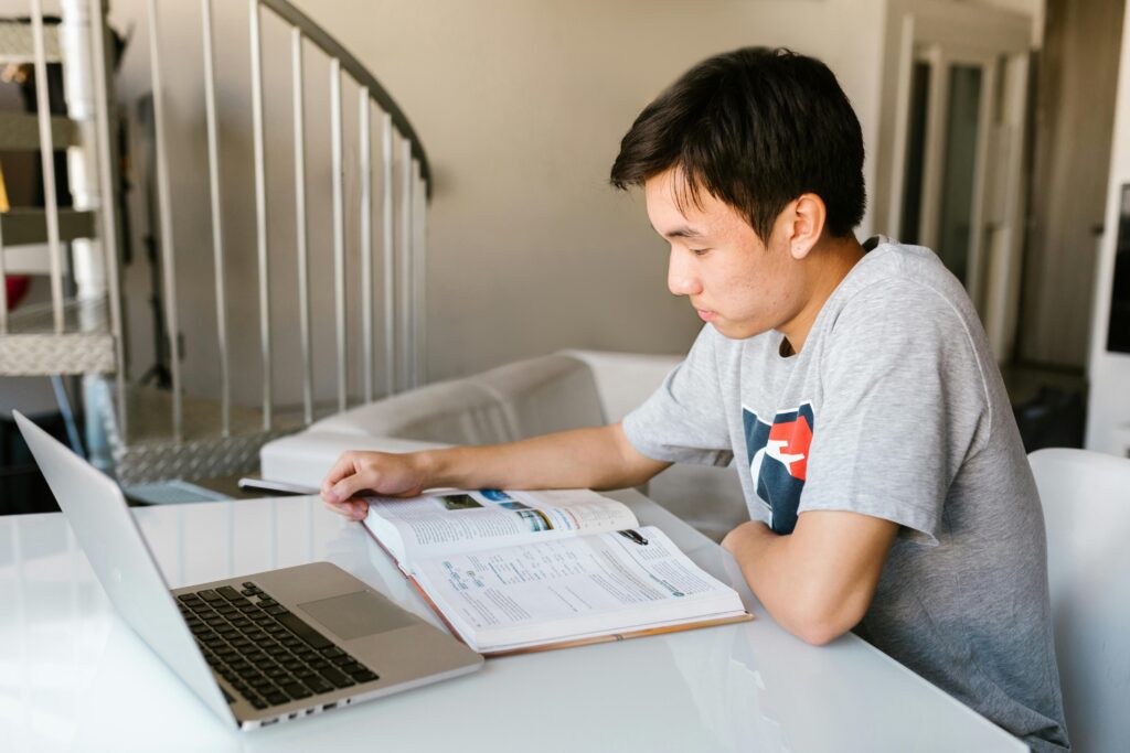 A young person studies at a table with an open book and a laptop in a bright room with a spiral staircase. | TCS