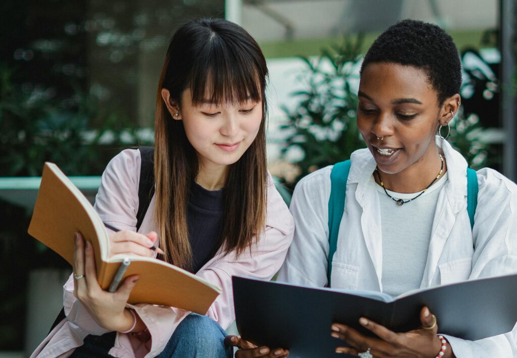 Two students sit together outdoors, reading and taking notes from exercise books, with rucksacks on their shoulders. | TCS