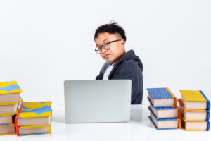 Boy with glasses sits at a desk with a laptop, surrounded by piles of books, against a white background. | TCS