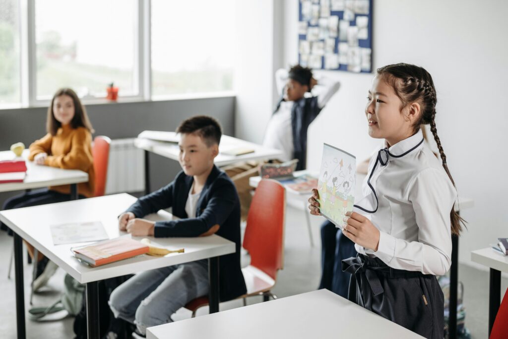 A girl stands and presents a picture book to her classmates in a bright, modern classroom. | TCS
