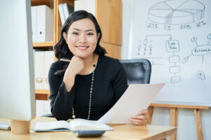 Businesswoman smiling at her desk, holding papers, with a notebook and pie chart on whiteboard in the background. | TCS
