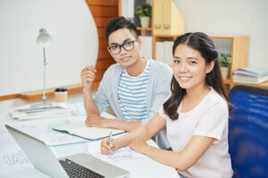 Two young adults work together at a desk with a laptop, papers, and notebooks, smiling at the camera. | TCS