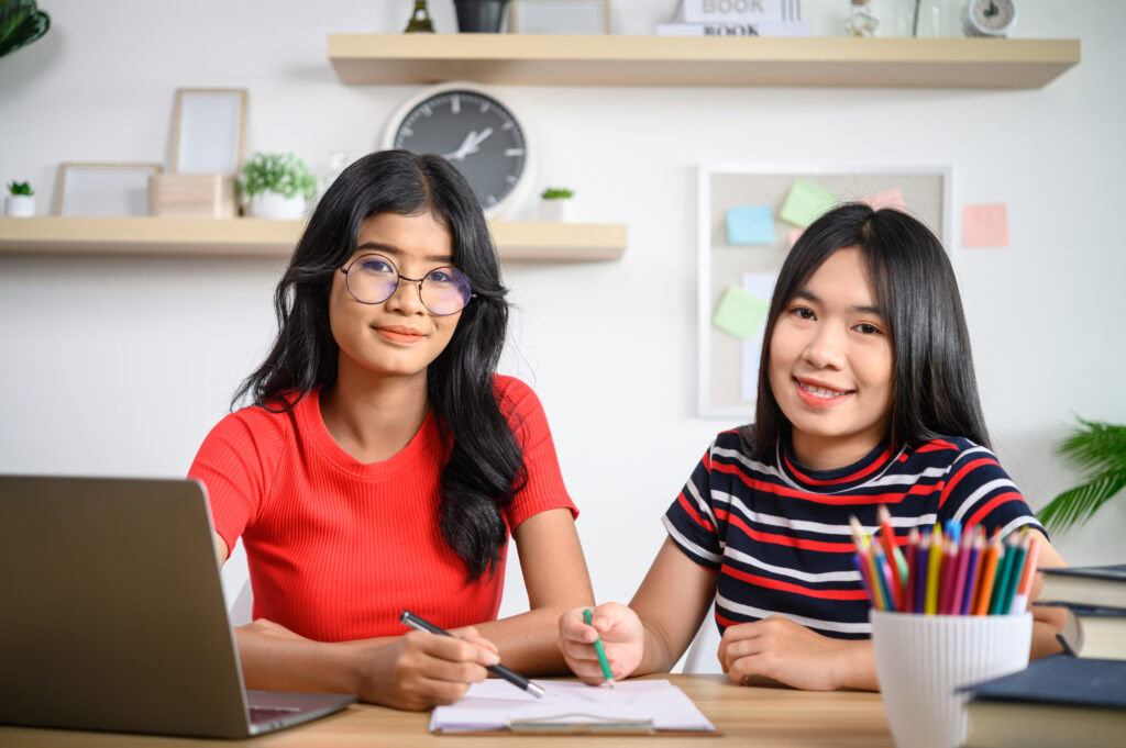 Two young women with long dark hair study at a desk with a laptop, notebooks, and coloured pencils. | TCS