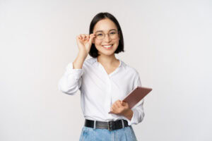 Smiling woman in glasses holding a tablet, wearing a white shirt and blue jeans, standing against a plain background. | TCS