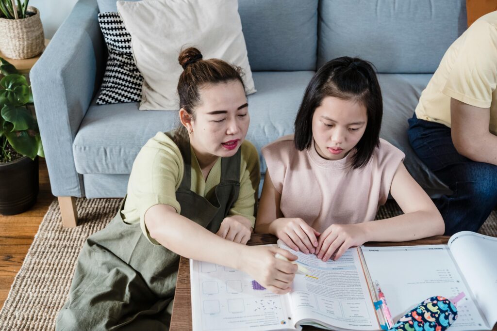 Two women sit at a table, studying and discussing an open textbook in a lounge setting. | TCS