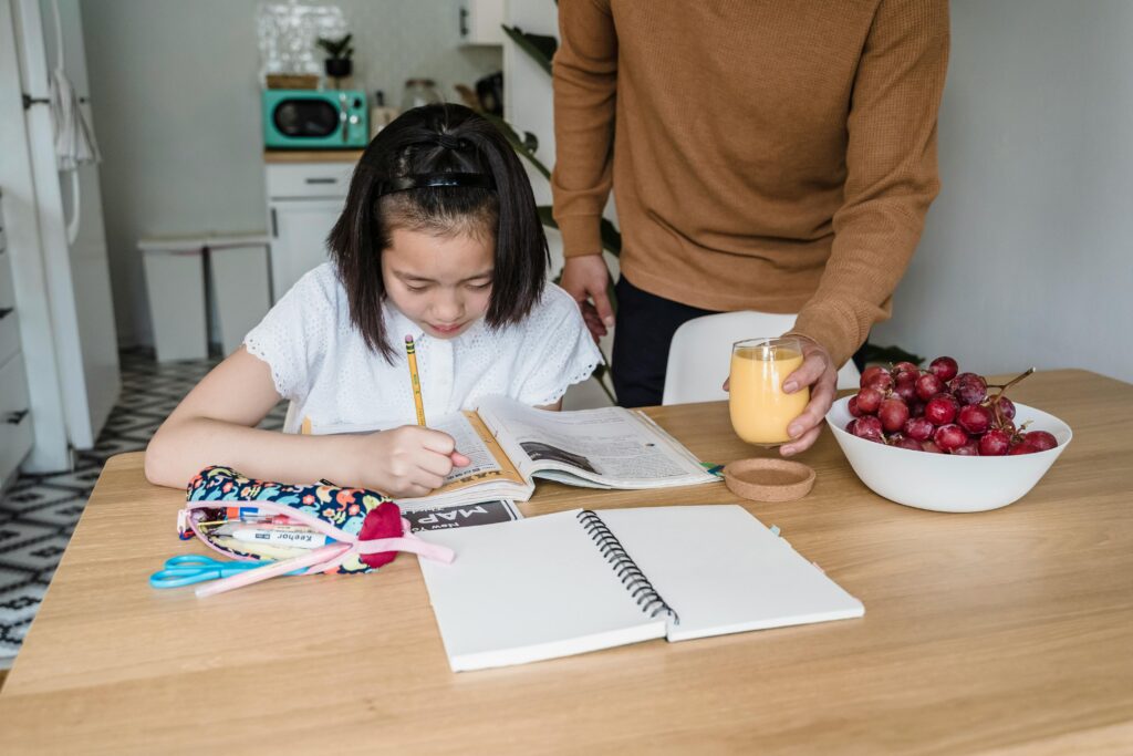 Young girl doing homework at a table while an adult hands her a glass of orange juice; grapes and stationery nearby. | TCS