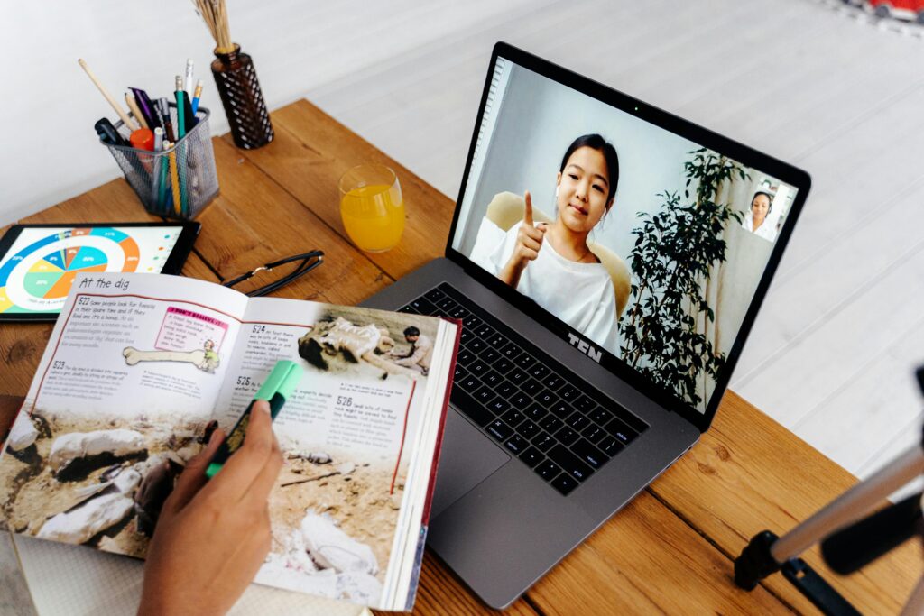 Person video chatting on laptop while studying a dinosaur book at a wooden desk with a drink, tablet, and stationery. | TCS