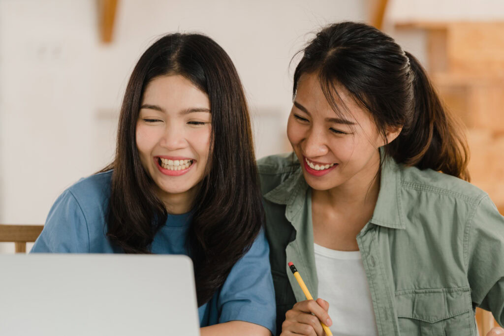 Two young women smiling at a laptop, discussing tuition methodology as one holds a pencil. | TCS