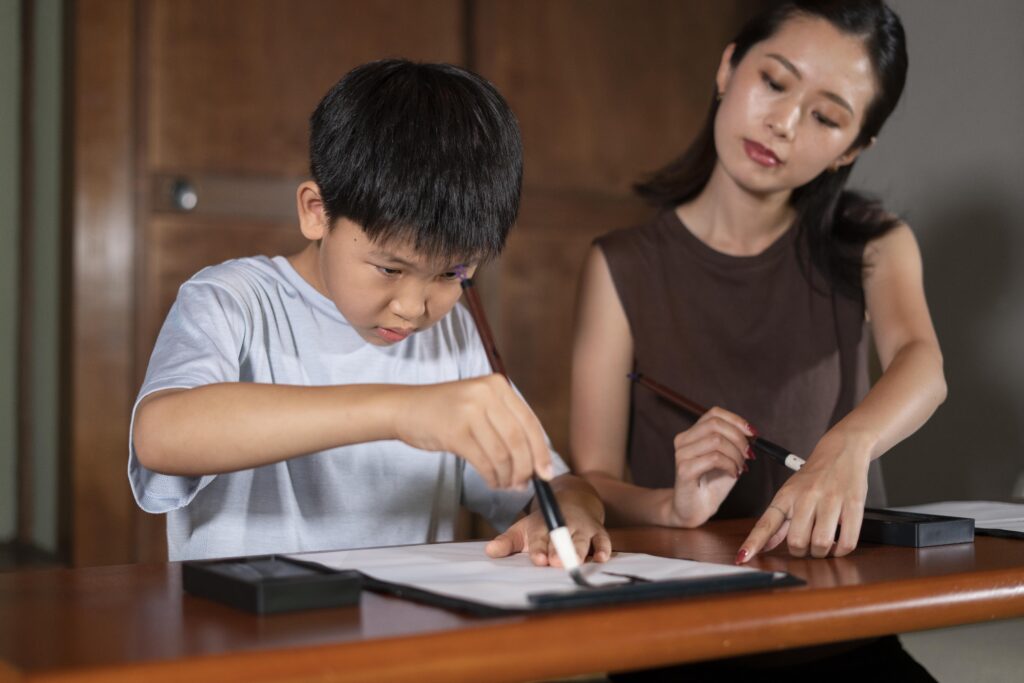 A boy and a woman practise calligraphy together at a table, both holding brushes and focussing on their work. | TCS