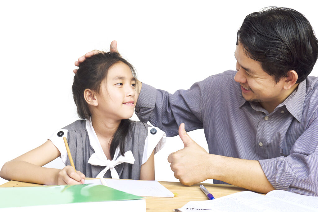 A man smiles and gives a thumbs up while patting a girl’s head as she does her homework at a table. | TCS