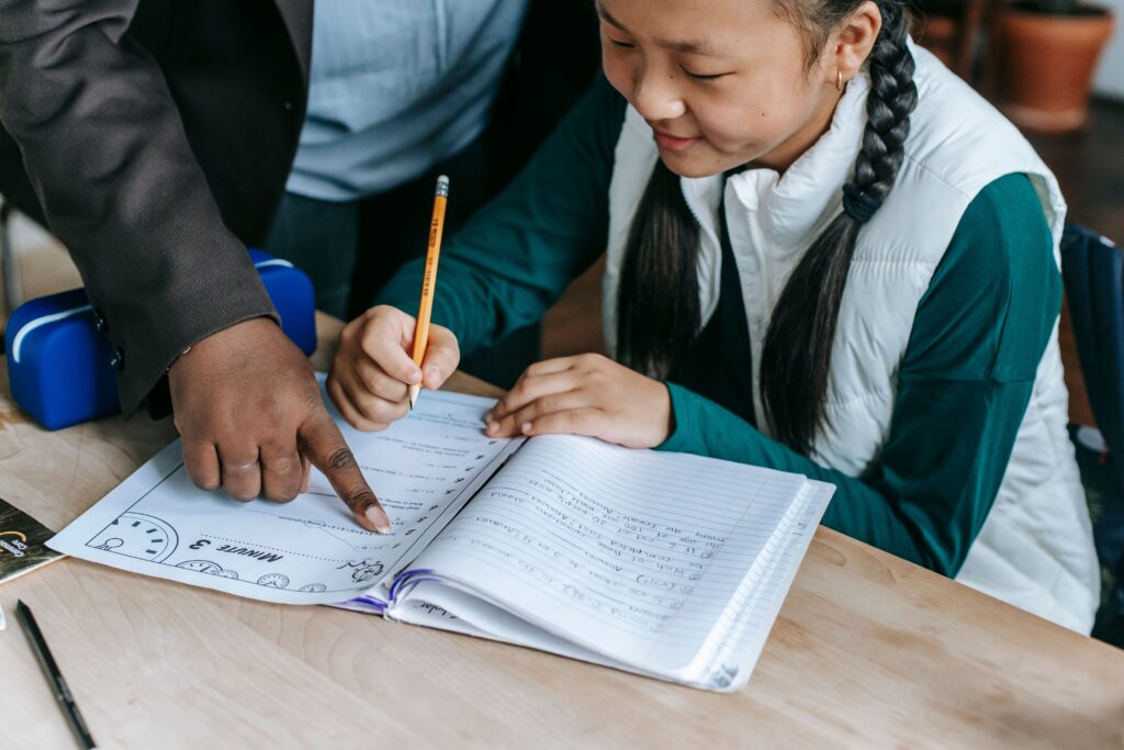 A student writes in a notebook while a teacher points to something on her open exercise book at a desk. | TCS