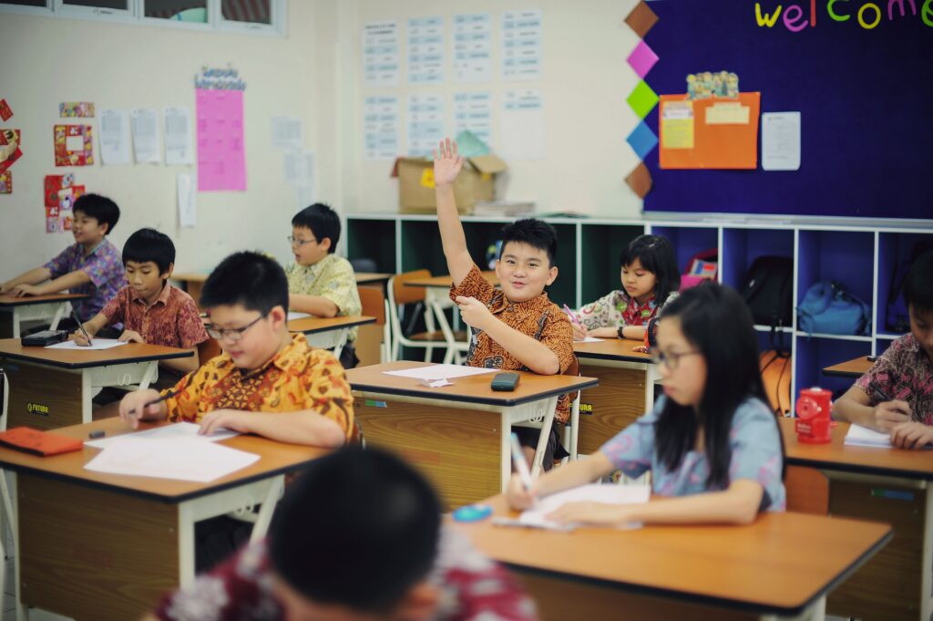 A boy raises his hand in a classroom whilst other pupils write at their desks. | TCS