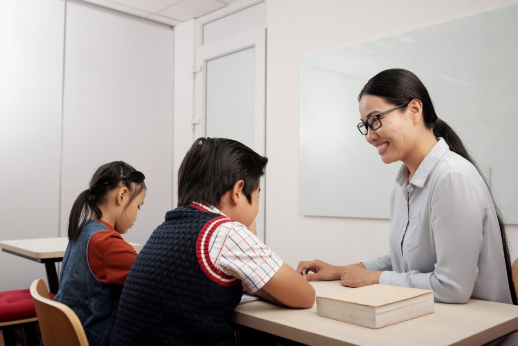 Smiling teacher uses effective teaching methodology to help two young pupils with their work at a classroom desk. | TCS