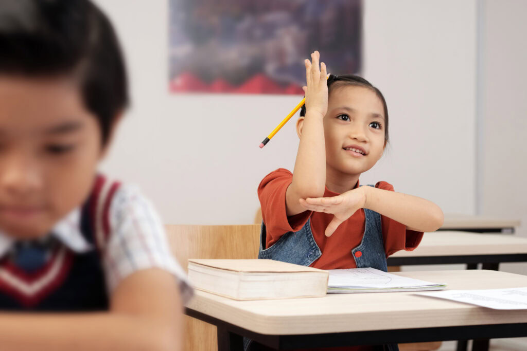 Smiling girl in a classroom raises her hand, holding a pencil, with books and papers on her desk. | TCS