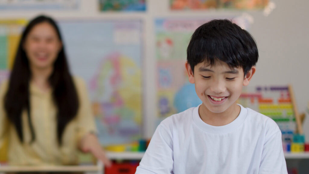 Smiling boy in a white shirt sits in a classroom, with a woman blurred in the background. | TCS