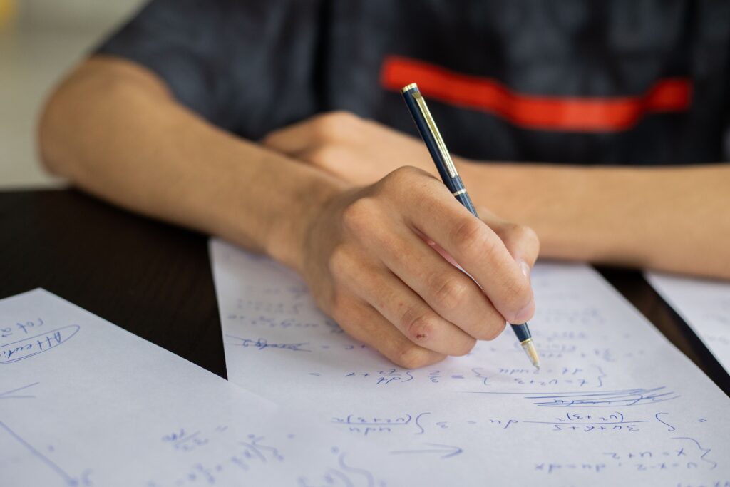 A person writes notes on white paper with a pen, surrounded by handwritten sheets on a dark table. | TCS