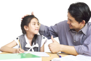 A man smiles, giving a thumbs-up, whilst patting a young girl doing homework at a desk. | TCS