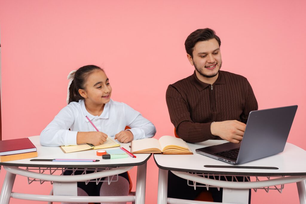 A girl writes in a notebook while a man beside her points at a laptop screen; both are smiling. | TCS