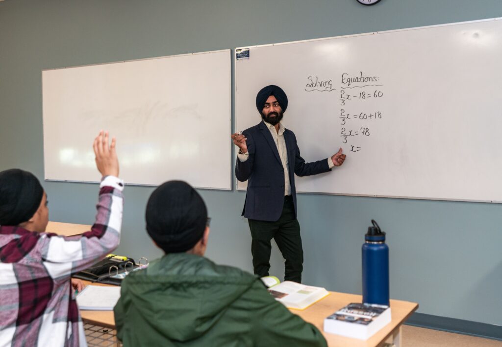 A teacher explains solving equations at a whiteboard as two pupils raise their hands in a classroom. | TCS
