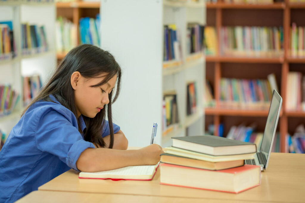 A student writes in a notebook at a desk with books and a laptop in a library setting. | TCS