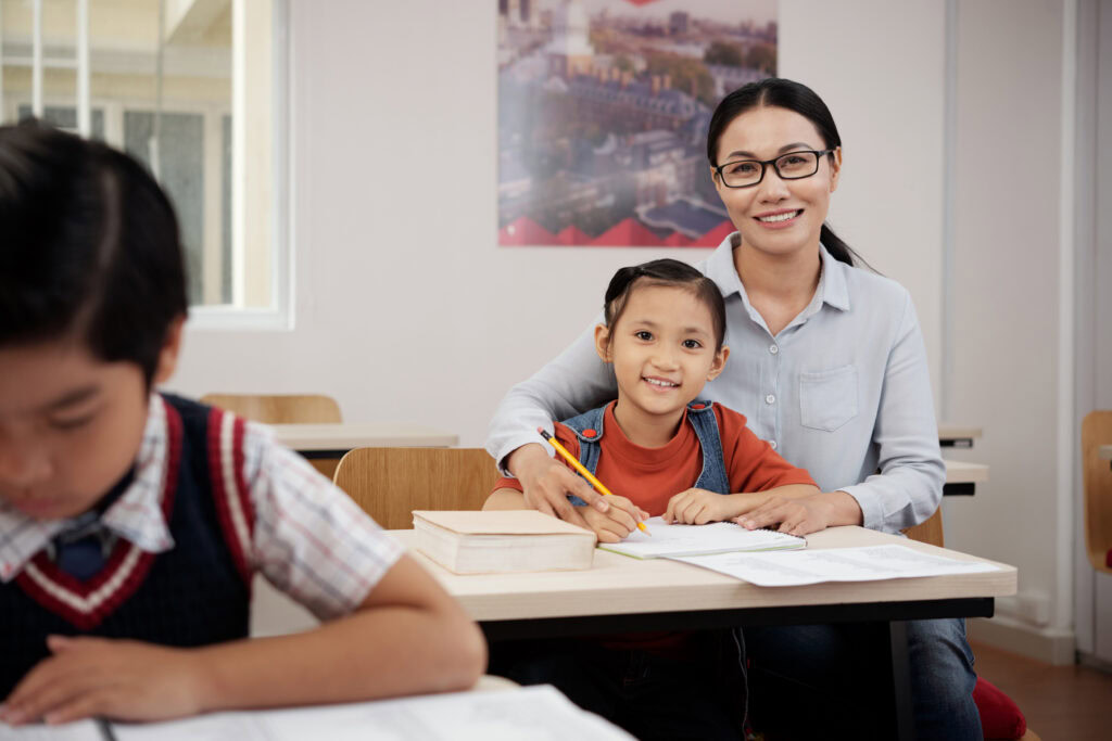 Smiling woman helps a young child with schoolwork at a classroom desk; another pupil sits in front. | TCS