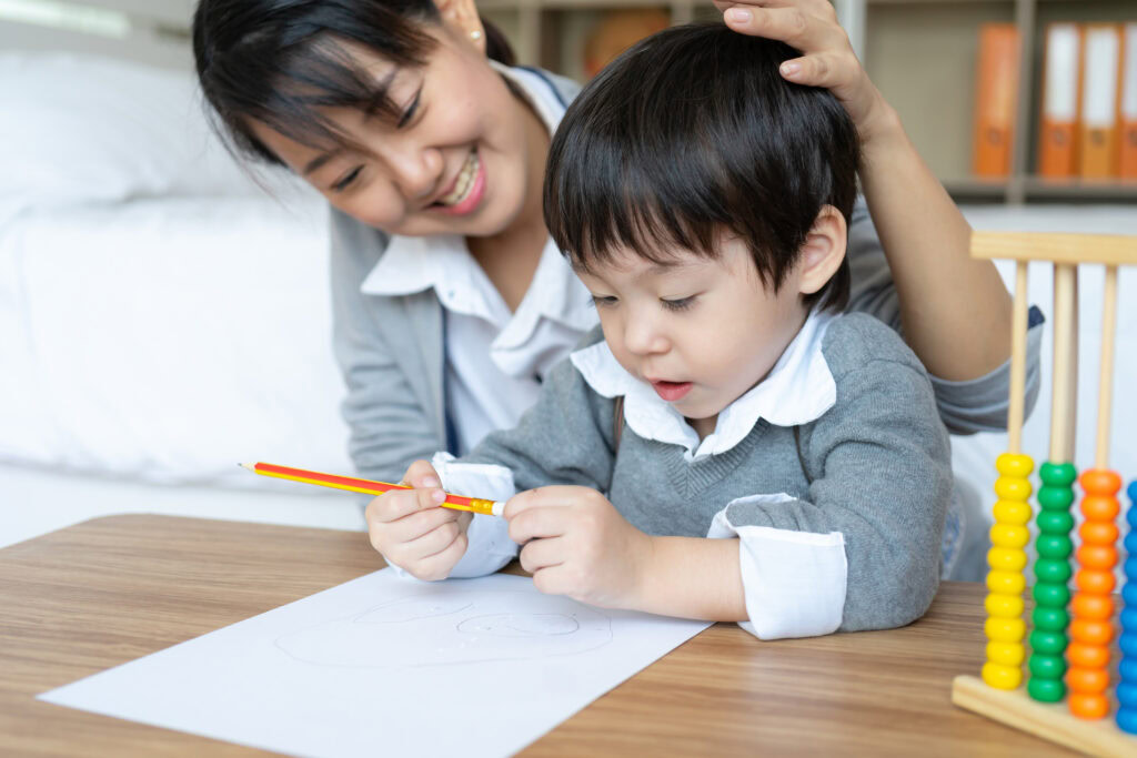 Smiling woman helps young boy draw with a pencil at a desk with an abacus and paper. | TCS