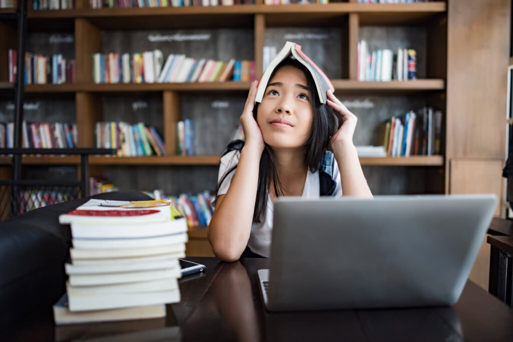 Young woman in a library with a book on her head, looking up, sitting at a table with a laptop and a stack of books. | TCS