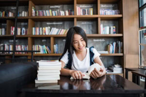 Young woman reading a book at a table with piles of books in a library or bookshop. | TCS