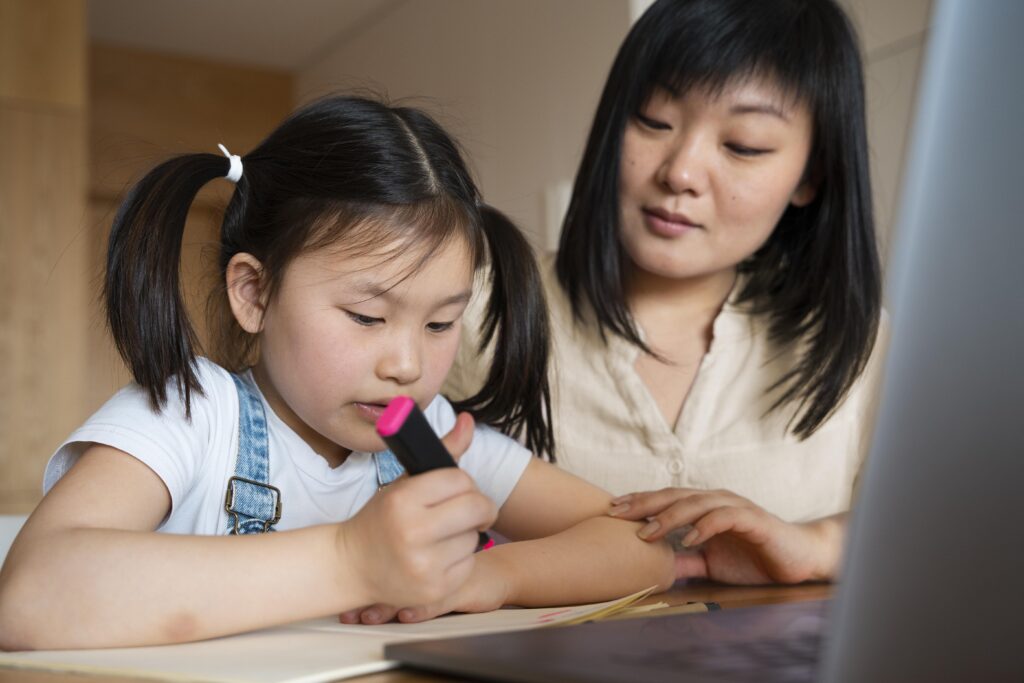 A woman helps a young girl with plaits as she writes in a notebook next to a laptop. | TCS
