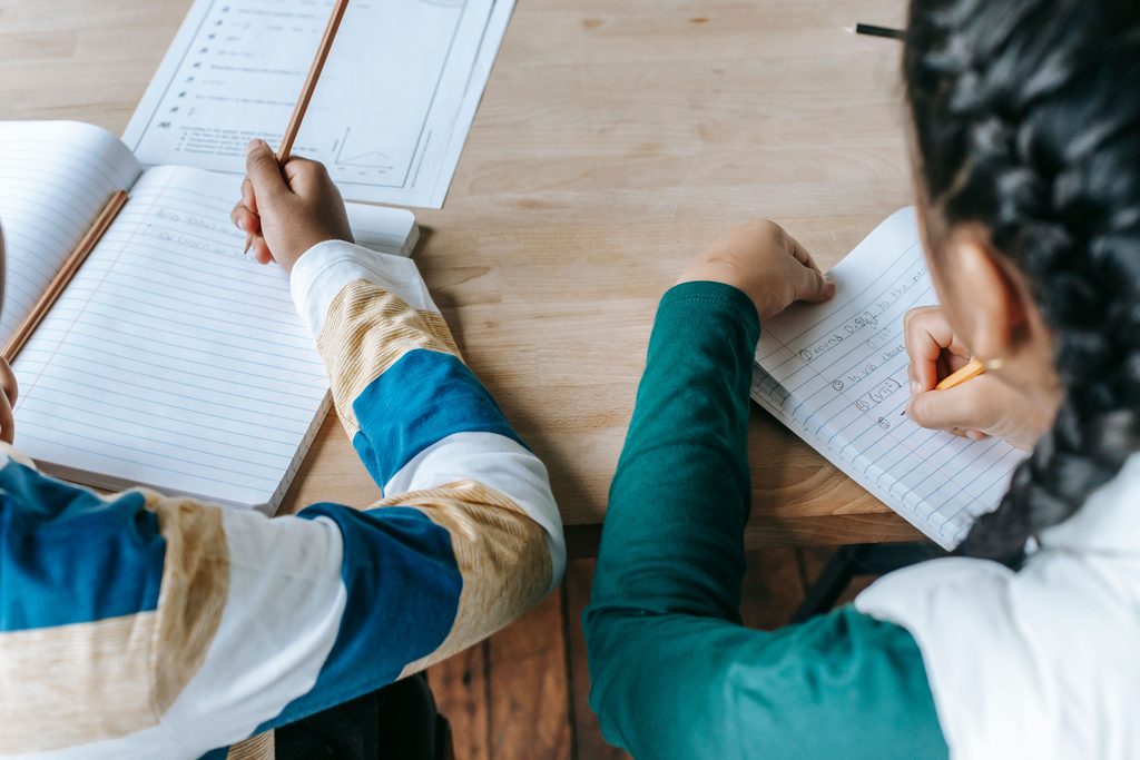 Two children sit at a table writing in exercise books, seen from above, with worksheets and pencils. | TCS