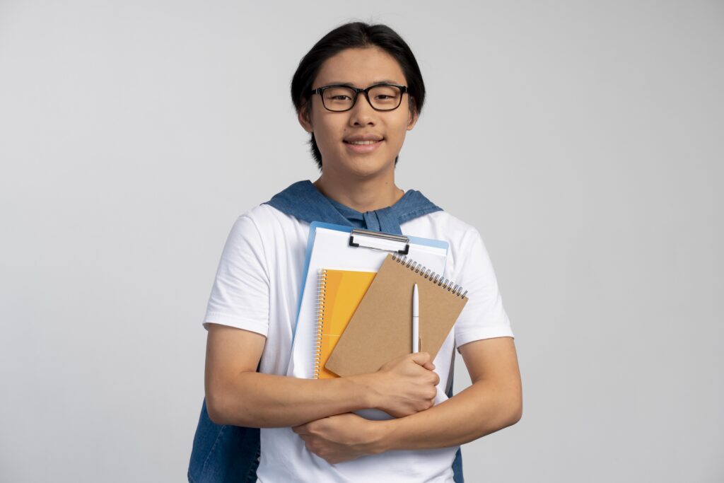 Smiling student with glasses holding notebooks, clipboard, and pen, wearing a white shirt and denim jacket over shoulders. | TCS