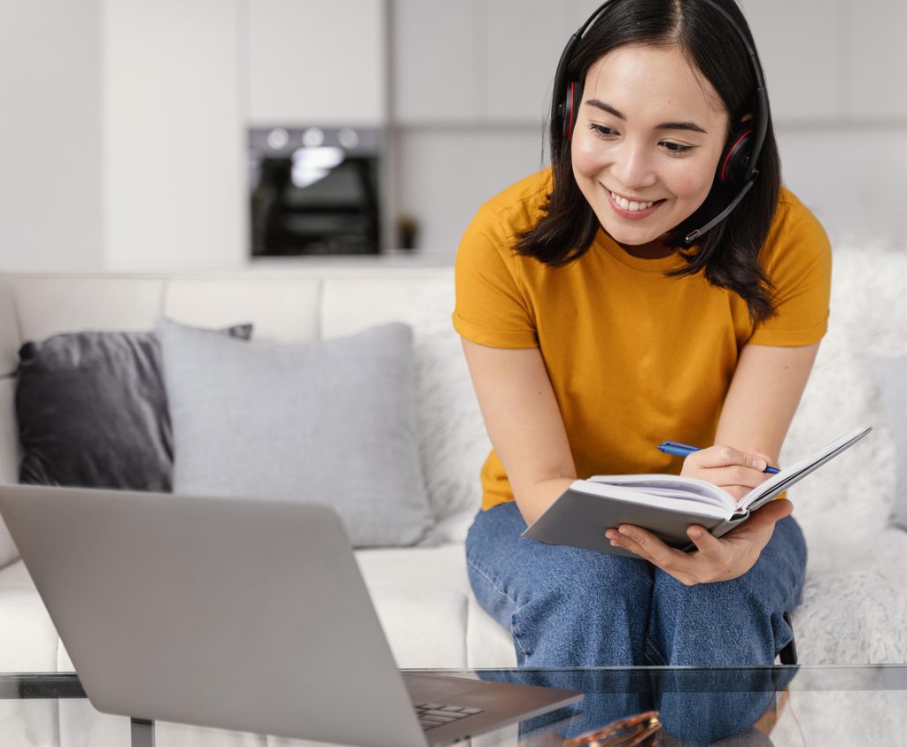 Woman in headset smiles, takes notes in notebook whilst watching a laptop on a glass table in a living room. | TCS