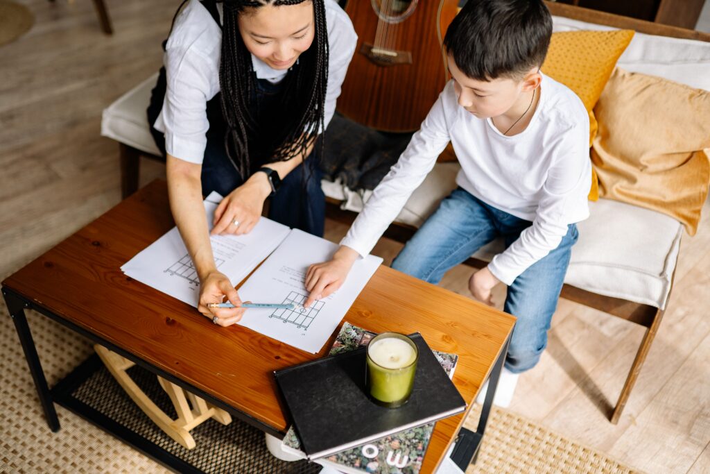 Woman offering emotional support as she helps a boy with his worksheet at a wooden table filled with study materials. | TCS