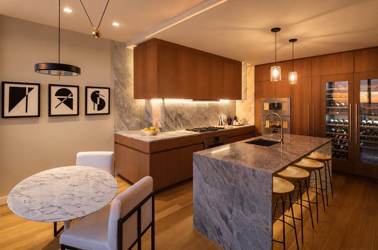 Luxury custom kitchen in NYC featuring warm wood cabinetry, natural stone countertops, waterfall island, integrated appliances, wine storage, and modern lighting by Urban Homes.