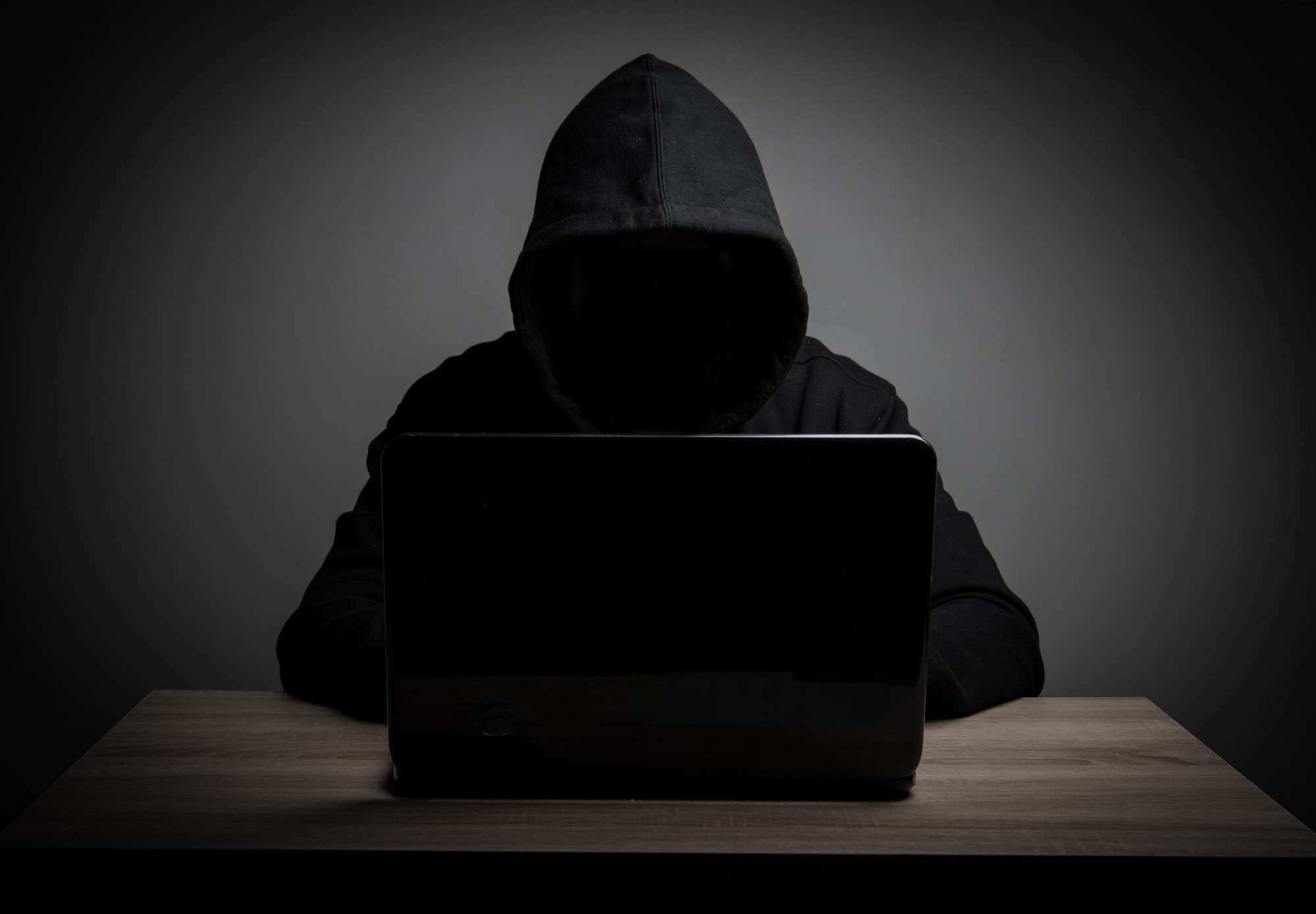 Sötétben ülő, kapucnis hálózati bűnöző laptop előtt.