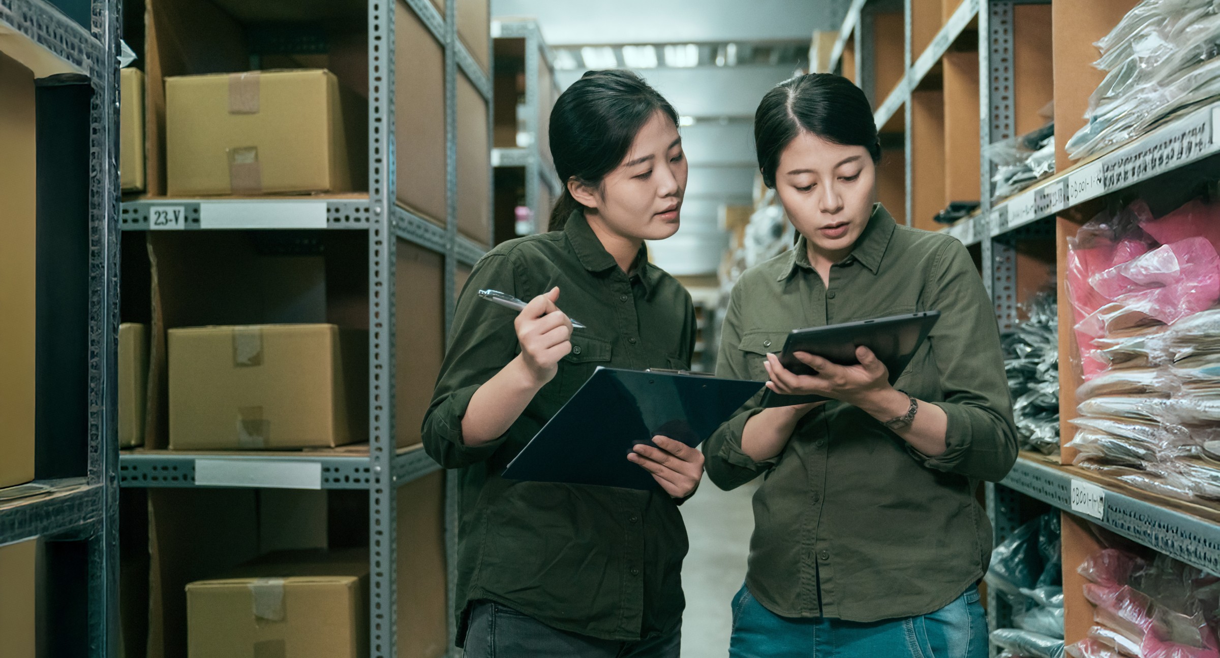 Two female employees in a warehouse, using a clipboard and a tablet to manage inventory, representing efficient uniform program management and uniform consultation services.