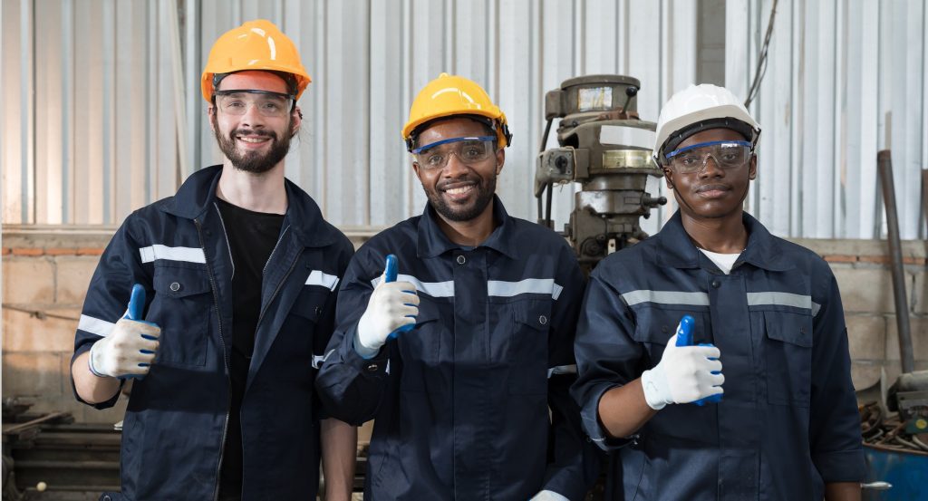 A diverse team of industrial workers in a factory, wearing professional high-quality uniforms and safety gear, giving a thumbs-up to represent employee satisfaction with their durable workwear.