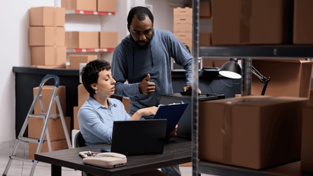 Warehouse employees reviewing a uniform order on a laptop in a distribution area