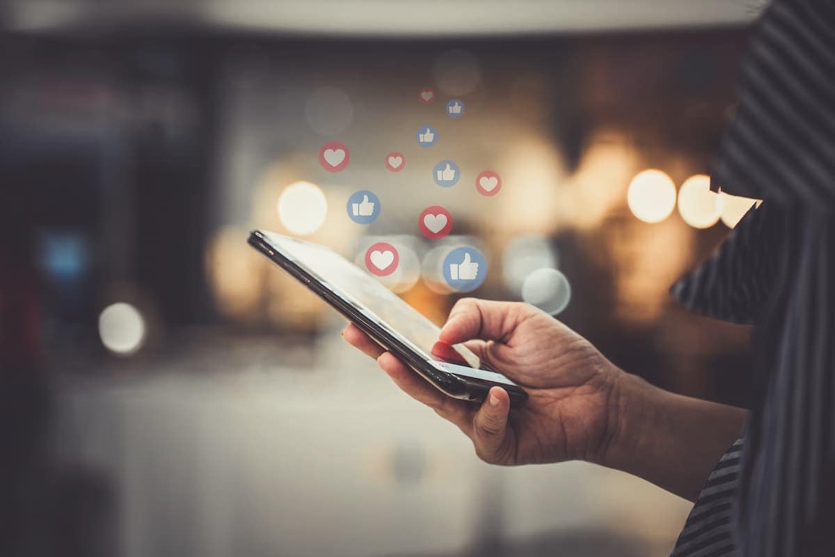 A phone being held in a a persons hand while illustrations representing social media likes float out of it