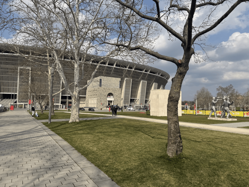 The exterior of Puskás Aréna seen through trees from the surrounding park.