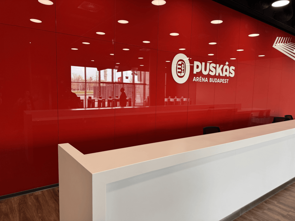 A white reception counter in front of a red Puskás Aréna wall.