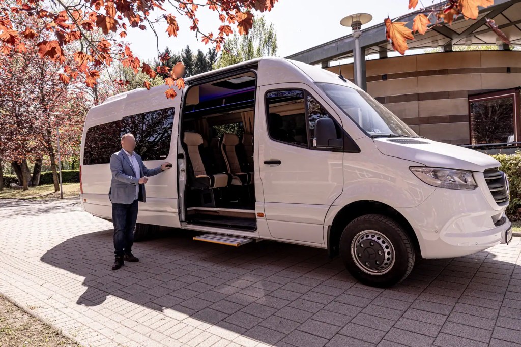 White Mercedes-Benz Sprinter minibus with its passenger door open and a chauffeur standing beside it