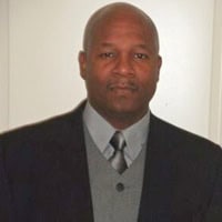 A man wearing a dark suit, gray sweater vest, and tie stands in front of a plain light background, looking at the camera.