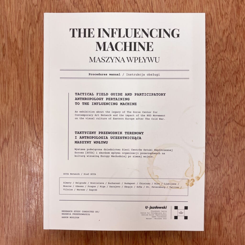 Photograph of a white paper titled 'The Influencing Machine' with text in English and Polish, placed on a wooden surface.
