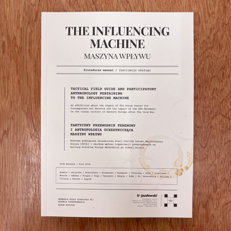 Photograph of a white paper titled 'The Influencing Machine' with text in English and Polish, placed on a wooden surface.