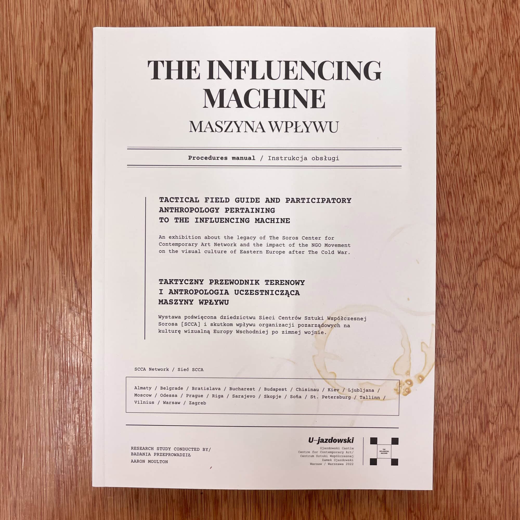 Photograph of a white paper titled 'The Influencing Machine' with text in English and Polish, placed on a wooden surface.